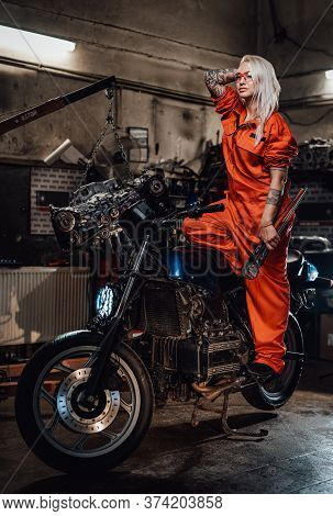 Tattooed Hipster Girl In Orange Overalls Hold A Big Wrench And Posing For A Camera While Standing On