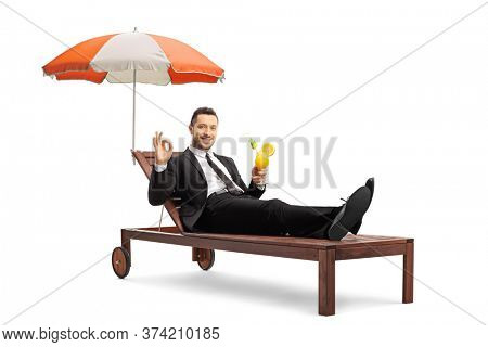 Businessman on a sunbed under umbrella holding a cocktail and gesturing an ok sign isolated on white background