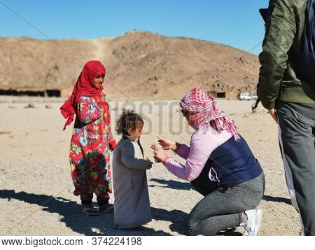 Bedouin Village In Desert, Egypt - February 2020: Tourists And Bedouins Kids, Middle View. Woman Giv