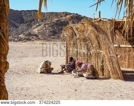 Bedouin Village In Desert, Egypt - February 2020: Four Bedouins Sitting Near Yellow Reed Shed, Mediu