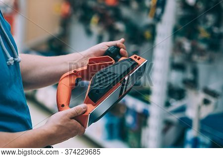 A Man Holds In His Hand An Electric Planer For A Tree. A Buyer In A Hardware Store Selects A Product