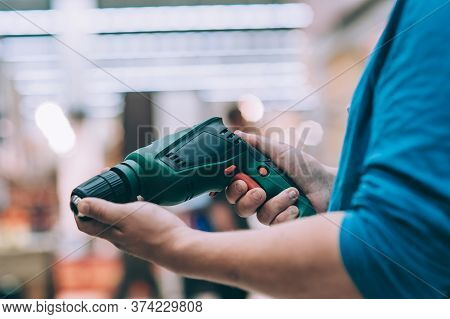 A Man Holds An Electric Drill In His Hand For Drilling. A Buyer In A Hardware Store Selects A Produc