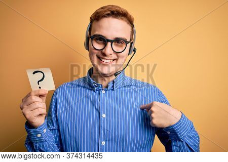 Redhead call center agent man working using headset holding reminder with question mark pointing finger to one self smiling happy and proud