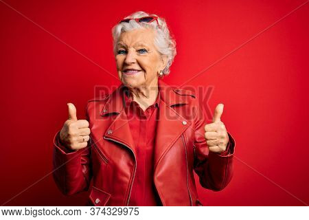 Senior beautiful grey-haired woman wearing casual red jacket and sunglasses success sign doing positive gesture with hand, thumbs up smiling and happy. Cheerful expression and winner gesture.