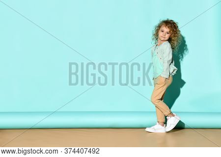 Pretty Curly Little Girl Standing Near The Wall With Hands In Pockets And Calm Serious Face. Full Le