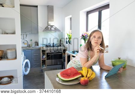 Shocked Young Woman Using Tablet Near Saucepan On Counter While Cooking In Her Kitchen, Leaning On T