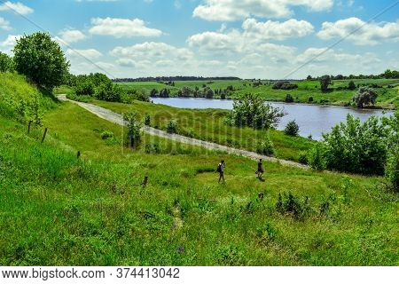 Two Male Tourists Walk Along A Path Among A Spring-summer Green Meadow Against A Pond On A Sunny Day