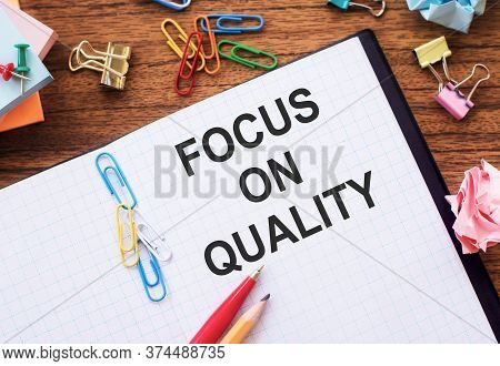 Notebook With Text Focus On Quality On A Table With Sticks, Colored Paper Clips And Wrinkled Sheets.