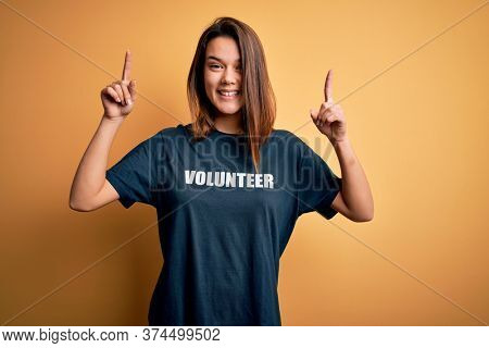 Young beautiful brunette girl doing volunteering wearing t-shirt with volunteer message word smiling amazed and surprised and pointing up with fingers and raised arms.