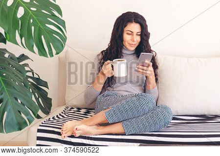 Caucasian Woman Using Smartphone While Relaxed Drinks Morning Coffee Sitting Comfortably On The Couc