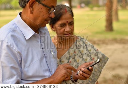 Senior Indian Bengali Couple In Park Looking At Their Smart Phone And Smiling In A Park In New Delhi