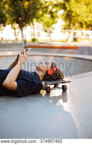 Young Skater In Orange Headphones Lying On Skateboard While Dreamily Using Cellphone Spending Time A