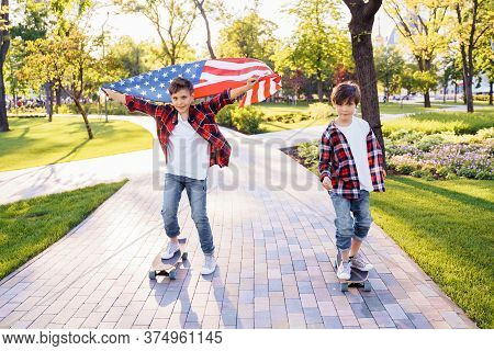 Two Cute Boys Brothers Riding Skateboards In A Public Park On A Sunny Afternoon. U.s. Independence D