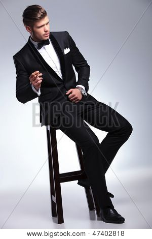 elegant ung mode manden i smoking sidder på en stol og ryge mens du kigger fra camer