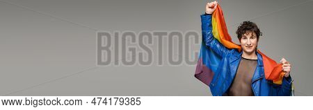 Happy Pangender Person In Blue Leather Jacket Posing With Rainbow Flag Isolated On Grey, Banner.