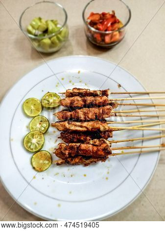 A Plate Of Chicken Satay Served With Lime, A Glass Of Pickles And Sambal Matah In The Room, Their Vi