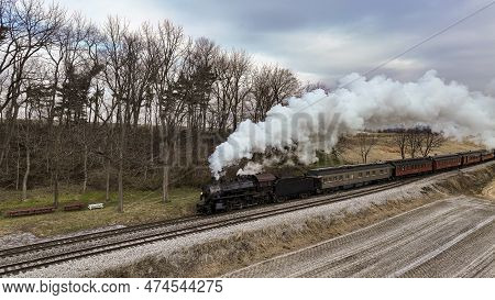 A Drone View Of A Steam Locomotive Approaching Traveling Thru Fields And Meadows, Blowing White Smok
