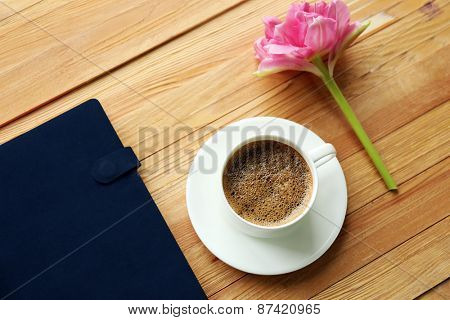 Cup of coffee with notebook and fresh flower on wooden table background