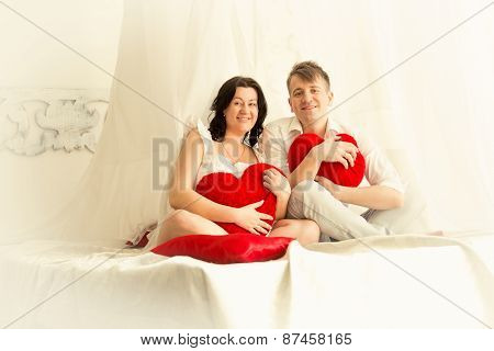 Toned Photo Of Pregnant Couple Posing On Big Bed With Baldachin