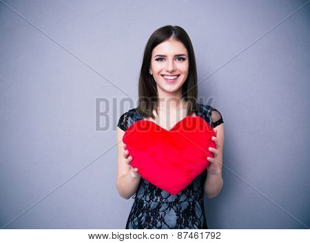 Smiling beautiful woman in dress holding red heart over gray background and looking at camera
