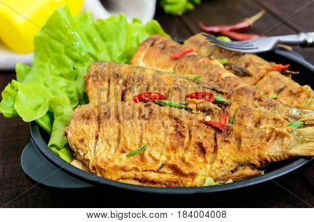 Fried fish carp (sazan) on a cast-iron frying pan with lettuce leaves on a dark wooden background. Close up