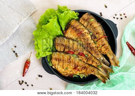Fried fish carp (sazan) on a cast-iron frying pan with lettuce leaves on a light background. The top view.