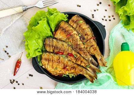 Fried fish carp (sazan) on a cast-iron frying pan with lettuce leaves on a light background. The top view.
