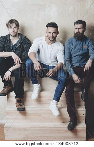 Three bearded men young friends or people with different emotions sitting on wooden stairs on beige wall. Handsome caucasian models with stylish hair in casual clothes. Beards and haircuts fashion