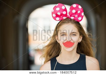 Decorative props on stick. Happy pretty girl or young woman teenager with long blond hair posing with cute mouse ears and paper red lips. Holiday birthday anniversary celebration. Fun party