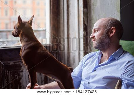 Dog And Its Owner Look Thoughtfully Out The Window, Watching The Street. Friendship Between A Dog An