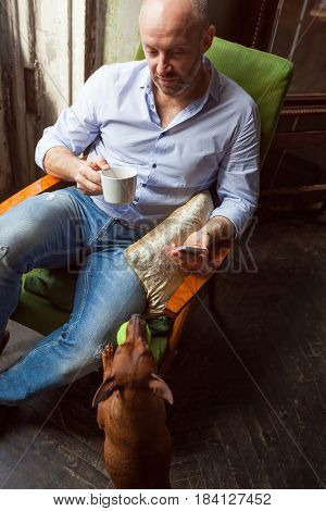 A Man In A Shirt Drinks A Cup Of Tea, And His Dog Invites Him To Play Ball Or Go For A Walk.