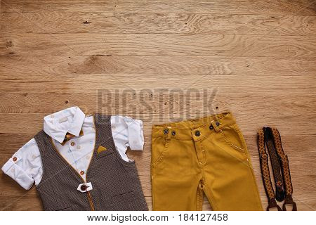 Top view photo of the boy's clothes with accessories on the wooden background. White shirt with print, brown vest, bowtie, brown pants and suspenders. Cute clothes for little boys. Concept of the children's fashion.