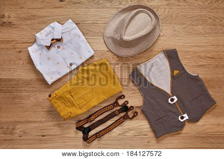 Top view photo of the boy's clothes with accessories on the wooden background. White shirt with print, brown vest, bowtie, brown pants, suspenders and hat. Cute clothes for little boys. Concept of the children's fashion.