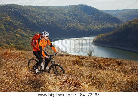 Cyclist in orange sporyive jacket riding the bike on the hill under river against beautiful mountain. Sportsman in the sportwear, helemet, sunglasses and with red backpack. Summer season. Healthy lifestyle.
