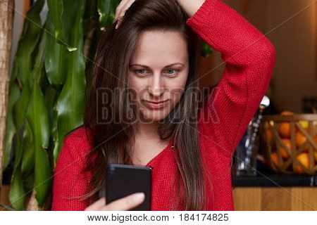 Close up shot of of beautiful young lady using her phone like a mirror or making selfie. Attractive woman is wearing red sweater sitting in a modern coffeehouse waiting for her lunch to come.
