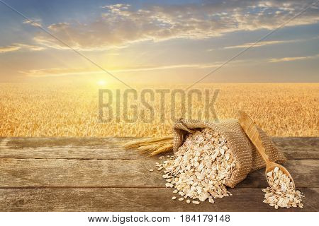 Oat flakes in bag. Still life scattered oatmeal, wooden spoon and ears on table with ripe cereal field on the background. Golden field on sunset. Healthy eating for diet and vegan