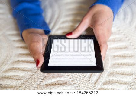 Sofia, Bulgaria - 14 April, 2017: Woman reading on her electronic book (e-book) on the bed at home. Hands only. Red nail polished fingers. Blue sleeves.