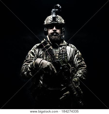 Bearded soldier in Combat Uniforms with weapon, plate carrier and combat helmet are on. Studio shot, dark background. Ruthless and fearless, he brings death to enemies, defending his country