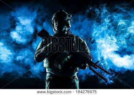 Army soldier in Combat Uniforms with machine gun, plate carrier and combat helmet are on. Studio contour silhouette shot, backlight, dark glowing smoke background