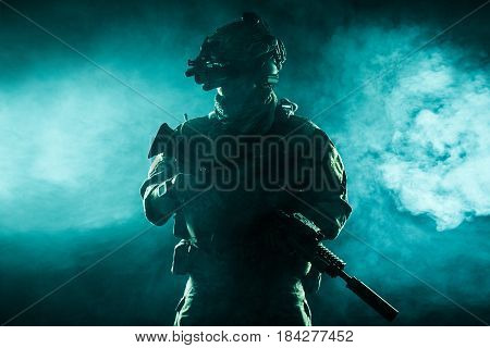 Army soldier in Combat Uniforms with assault rifle, plate carrier and combat helmet are on, Shemagh Kufiya scarf on his neck. Studio contour silhouette shot, backlight, dark glowing smoke background