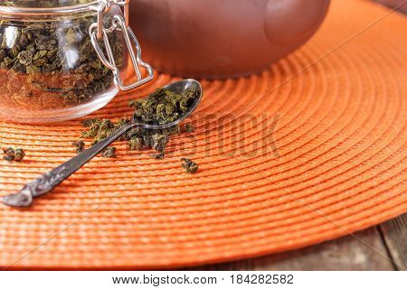 Dry leaves of green tea in a glass jar. Nearby tea leaves in a teaspoon and ceramic tea-things. Composition on a wattled napkin. The cover banks is open several tea leaves are scattered.