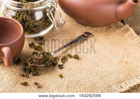 Dry leaves of green tea in a glass jar. Nearby tea leaves in a teaspoon and ceramic tea-things. Composition on a napkin from a sacking. The cover banks is open several tea leaves are scattered. Light background