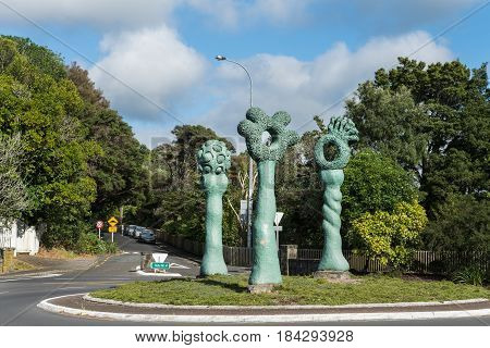 Auckland New Zealand - March 2 2017: The green obelisk-like Bush Markers statue on Titirangi roundabout. Green trees under blue sky with white clouds. Street with signs and cars.