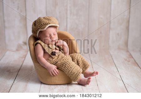 Nine day old newborn baby boy wearing a crocheted little man suit with newsboy cap and bowtie. He is sleeping in a tiny chair with a fist under his chin.