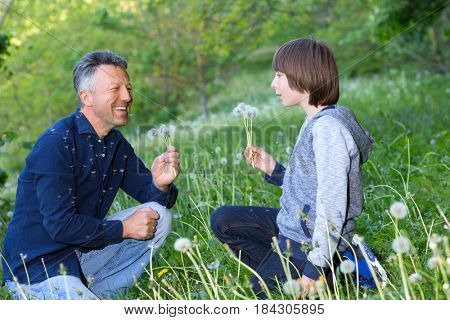 Man with his son blowing dandelions over blurred green grass, summer nature outdoor