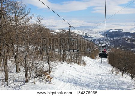 People ride in chairs of cableway among trees at winter day in Cahkadzor resort in mountains, Hrazdan town far away
