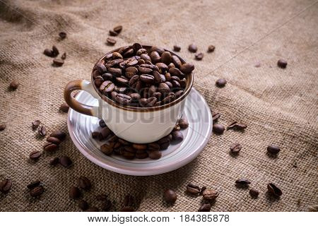 White Cup full of roasted coffee beans on rustic old cloth.
