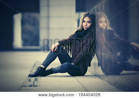 Happy young woman sitting at the wall in city street. Stylish fashion model in leather fringe suede jacket and blue jeans