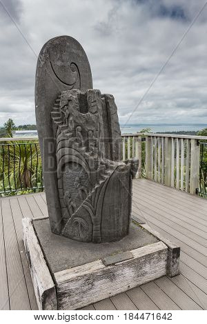 Auckland New Zealand - March 2 2017: Gray stone sculpture at the visitor center balcony celebrating the people who work to protect the Waitakere Ranges Park. Cloudscape and water of bay in back.