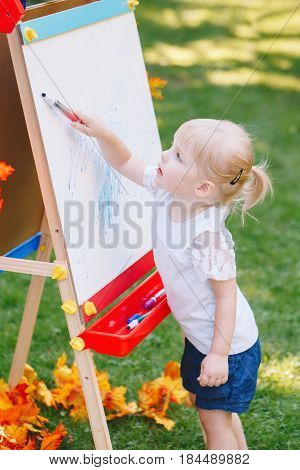 White Caucasian toddler child kid girl standing outside in summer autumn park drawing on easel with markers looking away playing studying learning back to school concept